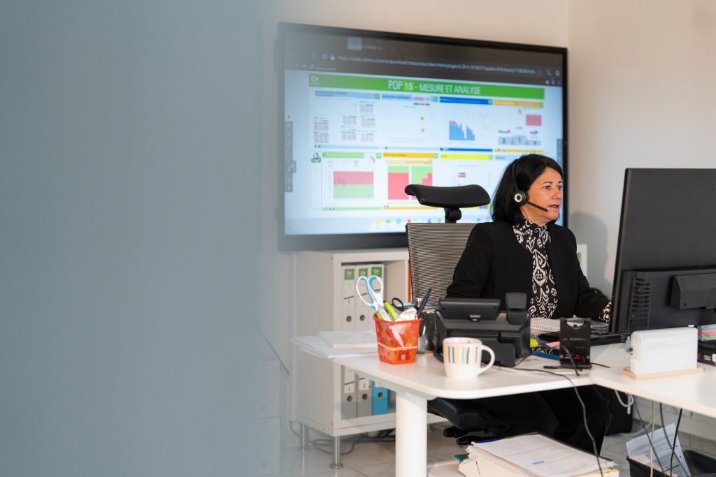 Femme utilisant un ordinateur avec un casque, devant un grand écran iObeya affichant un tableau de bord de gestion de la performance.